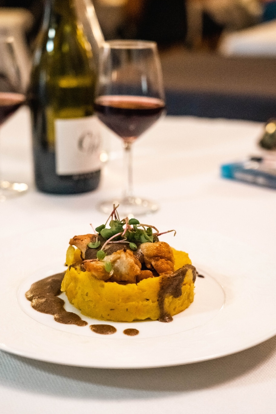 Plat gastronomique de morceaux de viande sur purée jaune (citrouille/potiron), sauce brune, et jeunes pousses, servi avec du vin rouge en arrière-plan.