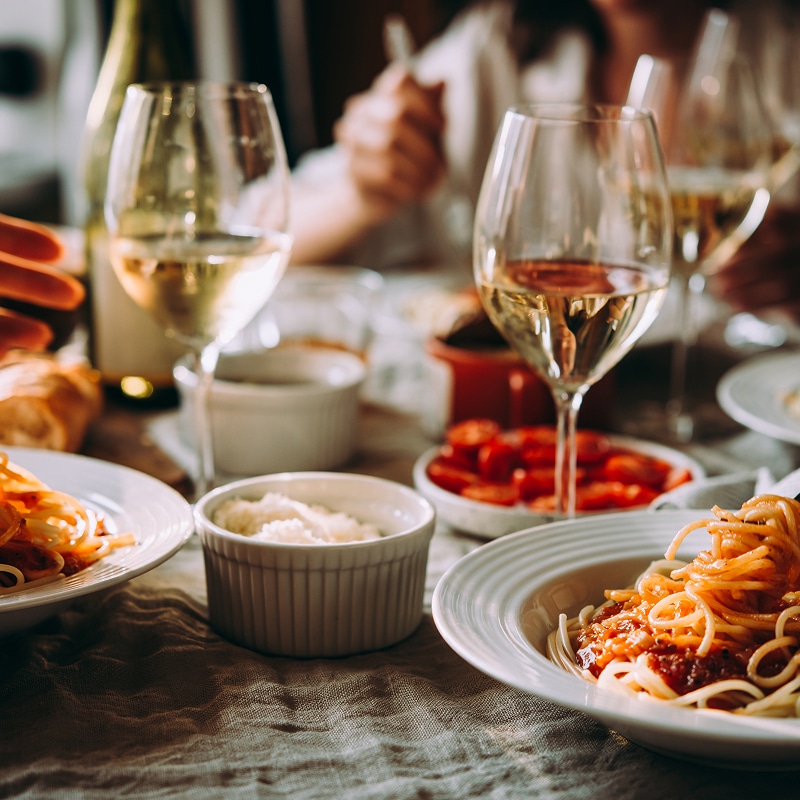 Dîner italien convivial : pâtes, vin blanc et amis Dîner italien convivial avec spaghettis à la sauce tomate, parmesan râpé et vin blanc dans des verres à pied.